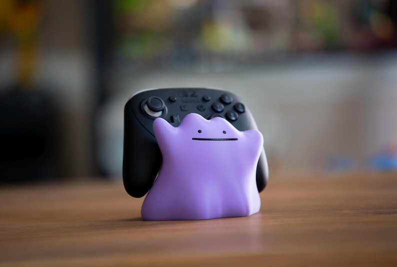 May include: A black video game controller with a purple Ditto-shaped stand. The controller has multiple buttons and joysticks. The Ditto stand is a light purple color with a simple face.