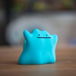 May include: A light blue Ditto figurine with a simple black line for a mouth and two small black dots for eyes. The figure is standing on a wooden surface. The background is blurred.