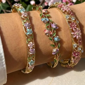 May include: Three gold-tone hinged bangle bracelets featuring intricate floral designs with enamel petals in shades of pink, blue, and purple, accented with green leaves and small clear crystals set within the metal filigree patterns.