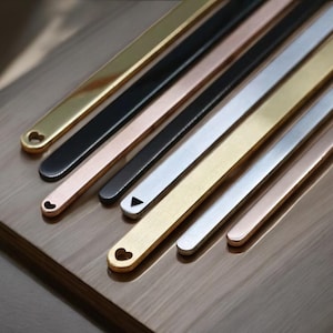 May include: A selection of metal bracelets in gold, black, silver, and rose gold. Some bracelets have heart or triangle cutouts. The bracelets are displayed on a wooden surface.