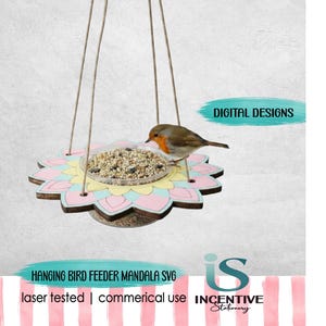 May include: A hanging bird feeder with a mandala-shaped wooden base painted in pastel pink, yellow, and blue. A clear circular tray filled with birdseed sits in the center. A small robin is perched on the edge. Text reads DIGITAL DESIGNS.