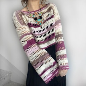May include: A crochet top in a striped pattern of white, purple, and brown. The long-sleeved top has a boat neck and flared sleeves. The open weave design adds a unique texture. A colorful statement necklace is also visible.