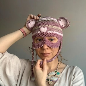 May include: A handmade crochet hat and mask set in shades of pink, lavender, and cream. The hat features bear ears and a heart-shaped appliqué. The mask has eye openings and beaded trim, with braided ties. A unique, whimsical accessory.
