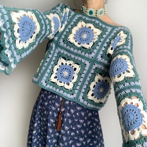 May include: Crocheted top in shades of teal, light blue, and white. The top features a square pattern with floral designs and bell sleeves. The garment is paired with a dark blue floral skirt.