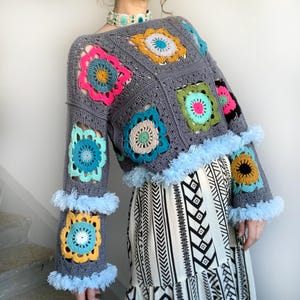 May include: A grey crochet sweater with colourful floral squares and light blue fringe trim. The sleeves and hem are adorned with the fringe. The sweater features a variety of colours, including pink, yellow, and teal.