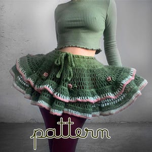 Puede incluir: Una falda de ganchillo verde con un diseño con volantes, con detalles en rosa y blanco. La falda tiene una cintura con cordón y está adornada con pequeñas flores rosas. La palabra "pattern" está escrita en cursiva en la parte inferior.