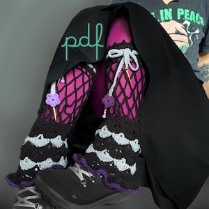 Puede incluir: Calentadores de piernas de ganchillo negros y grises con detalles en morado y azul claro. Los calentadores de piernas se usan sobre medias de rejilla rosas. La imagen también muestra una falda negra y zapatos negros.