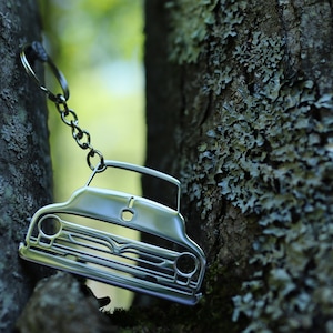 May include: Silver-colored car-shaped keychain hanging between tree branches. The keychain features a detailed outline of a classic car, including the grille, headlights, and roof. The chain is made of small, linked metal rings.