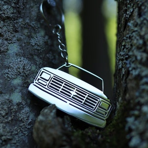 May include: A silver metal keychain shaped like the front of a pickup truck. The keychain has a detailed grille and headlights.