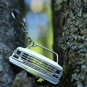 May include: A silver metal keychain shaped like the front of a vintage pickup truck. The keychain is hanging from a silver chain and is partially obscured by a tree trunk.