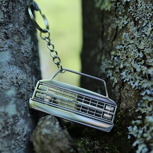 May include: A silver metal keychain shaped like the front of a pickup truck. The keychain has a detailed grille and headlights. The keychain is hanging from a silver chain.