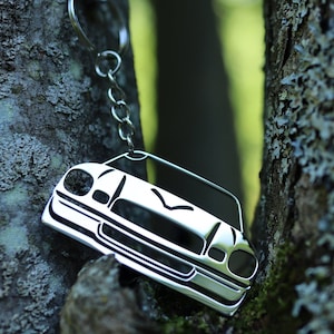 May include: A silver metal keychain shaped like a classic car. The keychain is hanging from a silver chain and is partially obscured by a tree trunk.