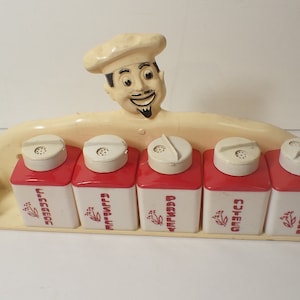 May include: A white plastic spice rack with a chef figurine on top. The rack holds five red and white spice containers with labels that read "Salt", "Pepper", "Garlic", "Oregano", and "Spice".