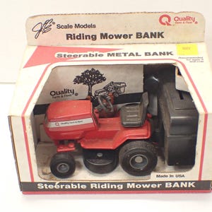 Puede incluir: Una alcancía de juguete de cortacésped rojo y negro con una rueda delantera orientable. La alcancía está en su caja original, que tiene las palabras "Scale Models Riding Mower BANK" y "Steerable METAL BANK" impresas en ella. La caja también tiene una imagen de un árbol y un pequeño cortacésped de juguete.