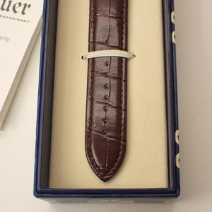 May include: A brown leather watch strap with a crocodile pattern is displayed inside a blue box with a cream-colored interior. A white elastic band secures the strap. The box also contains a white card with text and social media icons.
