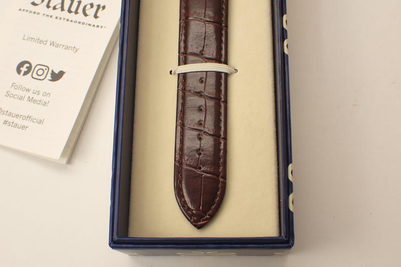 May include: A brown leather watch strap with a crocodile pattern is displayed inside a blue box with a cream-colored interior. A white elastic band secures the strap. The box also contains a white card with text and social media icons.