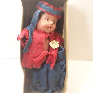 Puede incluir: Muñeca vintage en una caja de cartón. La muñeca viste un atuendo de fieltro rojo con detalles azules, incluyendo un sombrero y pantalones. La muñeca tiene una cara pintada con mejillas rosadas y está sentada.