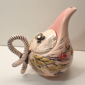 May include: A ceramic teapot with a pink spout and handle, and a light pink body. The body is decorated with black line drawings, green leaves, and red and yellow flowers. The handle is striped.