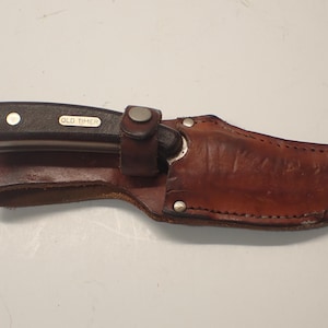 May include: A brown leather sheath containing a knife. The knife features a black handle with the text "OLD TIMER". The sheath is a darker brown and includes a strap to secure the knife.