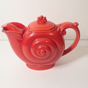 Puede incluir: Una tetera de cerámica roja con forma de caracol con un diseño de espiral. La tetera tiene una tapa con un adorno decorativo.