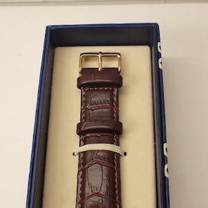 May include: A brown leather watch strap with a gold-colored buckle, presented in a blue box with a cream-colored interior. The strap has a textured, crocodile-like pattern and is secured with a white band.