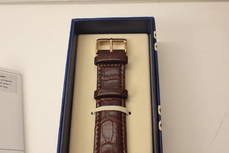May include: A brown leather watch strap with a gold-colored buckle, presented in a blue box with a cream-colored interior. The strap has a textured, crocodile-like pattern and is secured with a white band.