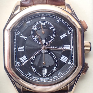 May include: A gold-toned wristwatch with a black face, featuring Roman numeral hour markers and multiple sub-dials. The watch has a brown leather band and the text "WATER RESISTANT 3ATM" is visible on the face.