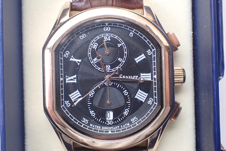 May include: A gold-toned wristwatch with a black face, featuring Roman numeral hour markers and multiple sub-dials. The watch has a brown leather band and the text "WATER RESISTANT 3ATM" is visible on the face.