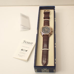 May include: A brown leather strap watch with a gold-colored case and a dark face, presented in a blue box. The watch has a rectangular face and is secured with a buckle. The brand name "Stauer" is visible on a card.