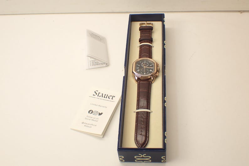 May include: A brown leather strap watch with a gold-colored case and a dark face, presented in a blue box. The watch has a rectangular face and is secured with a buckle. The brand name "Stauer" is visible on a card.