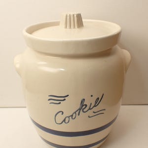 May include: Cream-colored ceramic cookie jar with a lid and two small handles. The jar has blue stripes around the base and the word "Cookie" in a handwritten font. The lid has a ribbed handle.