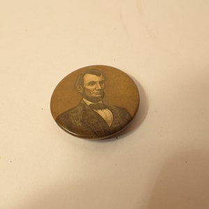 Puede incluir: Un botón redondo con fondo dorado y una imagen en blanco y negro de Abraham Lincoln.