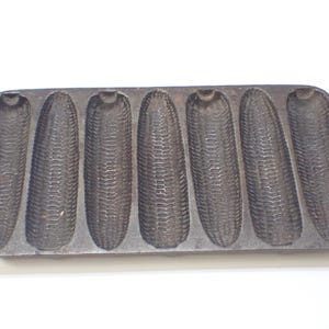 May include: A cast iron cornbread pan with eight individual moulds shaped like ears of corn. The pan is dark grey and has a rustic look.