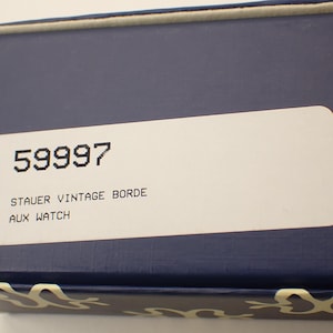 May include: A navy blue box with a white label. The label has the number "59997" printed in black, along with the text "STAUER VINTAGE BORDE AUX WATCH". The box has a decorative pattern along the bottom edge.