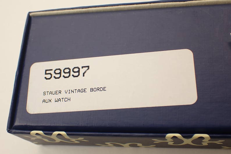 May include: A navy blue box with a white label. The label has the number "59997" printed in black, along with the text "STAUER VINTAGE BORDE AUX WATCH". The box has a decorative pattern along the bottom edge.