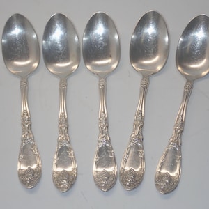 Puede incluir: Cinco cucharas de plata con mangos ornamentados y cuencos ovalados se disponen sobre una superficie gris claro. Las cucharas tienen un diseño detallado de flores y uvas en los mangos y cuencos, lo que sugiere un estilo vintage o antiguo.