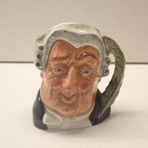 May include: A ceramic mug shaped like a person's head. The mug is painted to look like a judge with a white wig and a black robe. The handle is shaped like a green leaf.