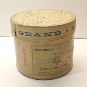 May include: A cylindrical, vintage cardboard container with the word "GRAND" in large, dark letters. The container has labels with text, including "Columbia Record" and "Grand Record No.". The overall color is tan with faded text and graphics.