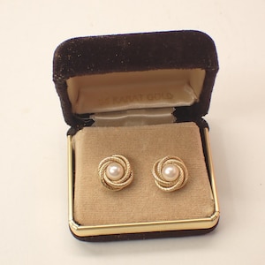 May include: A pair of gold stud earrings featuring a central white pearl surrounded by a textured, twisted rope design. The earrings are presented in a dark brown velvet jewelry box with a cream-colored interior lining and gold-tone trim.