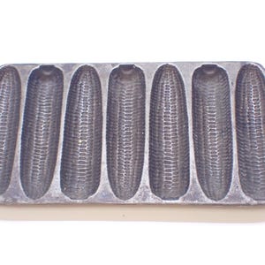 May include: A cast iron cornbread pan with eight individual moulds shaped like ears of corn. The pan is grey and has a rustic look.