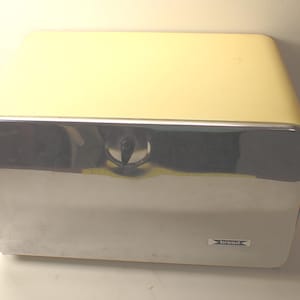 May include: A rectangular bread box with a pale yellow top and sides, featuring a reflective chrome front door. A small black knob is centered on the door for opening, and a white rectangular label with the word bread is attached to the lower right.