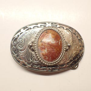 Puede incluir: Hebilla de cinturón ovalada plateada con un diseño floral grabado. El centro presenta una piedra ovalada con tonos naranja, marrón y crema. La hebilla tiene un estilo western clásico.