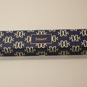 May include: A rectangular box with a navy blue background and a repeating gold pattern. The box has a gold-bordered label with the name "Stauer" in gold lettering. The box is likely for jewelry or a similar item.