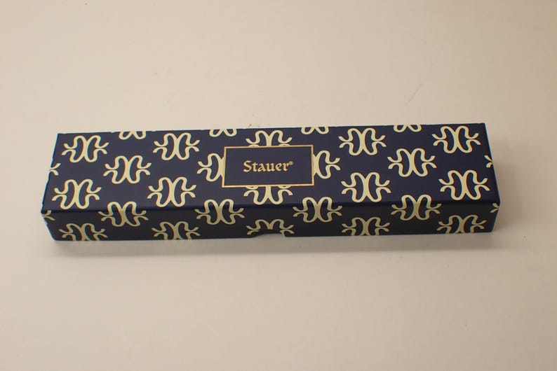 May include: A rectangular box with a navy blue background and a repeating gold pattern. The box has a gold-bordered label with the name "Stauer" in gold lettering. The box is likely for jewelry or a similar item.