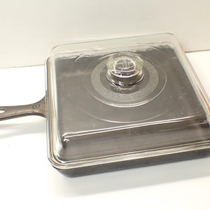 May include: Square cast iron skillet with a handle and a clear glass lid. The lid has a clear glass knob in the centre. The skillet is dark grey and has a handle on the left side. The lid is square and fits snugly on the skillet.