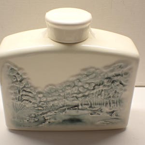 May include: Cream-colored ceramic container with a matching lid. The front features a landscape scene in shades of gray and blue, depicting trees and a body of water. The container has a rectangular shape.