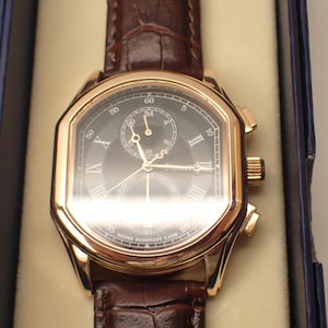 May include: A gold-toned wristwatch with a dark face and brown leather band. The watch features Roman numeral hour markers and a small subdial. The watch is presented in a blue box with a cream-colored interior.