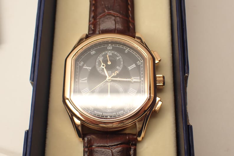 May include: A gold-toned wristwatch with a dark face and brown leather band. The watch features Roman numeral hour markers and a small subdial. The watch is presented in a blue box with a cream-colored interior.