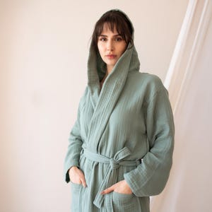 May include: A sage green hooded robe with a tie waist and pockets. The robe is made of a soft, textured fabric and has long sleeves. The model is wearing the robe.