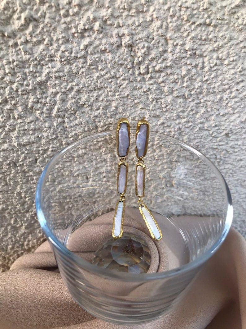 May include: A pair of drop earrings featuring three elongated, iridescent white stick pearls arranged vertically. Each pearl is encased in a thin, textured gold-toned metal frame, connected by small gold-toned jump rings to create a dangling effect.